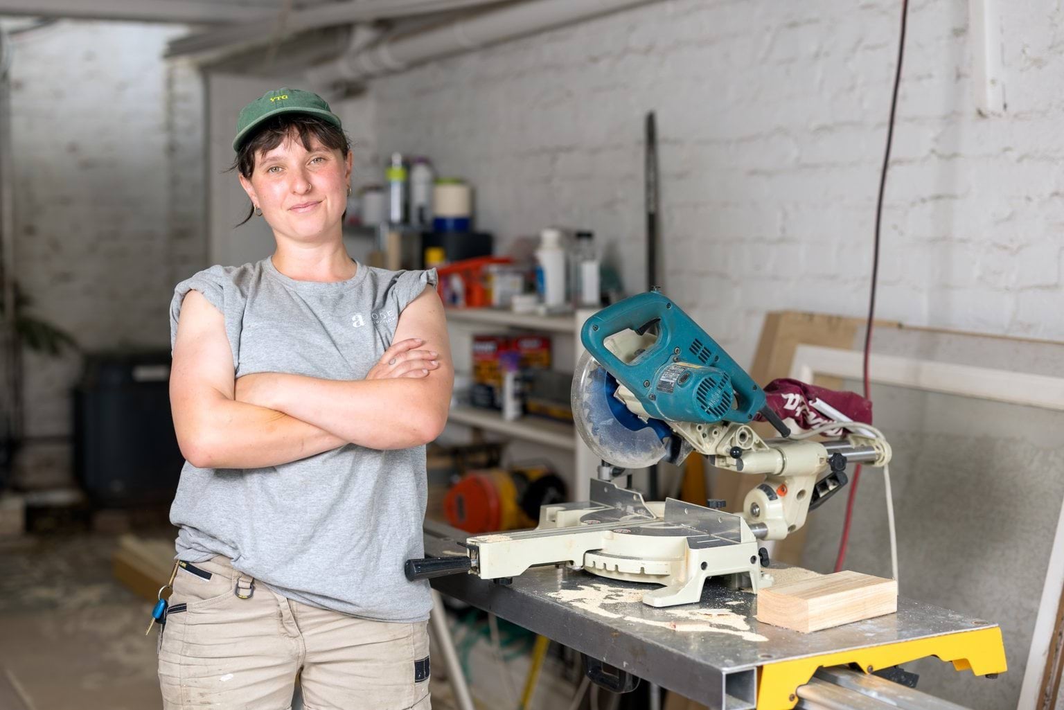 Tash is wearing a baseball cap and standing with arms folded in a workshop.