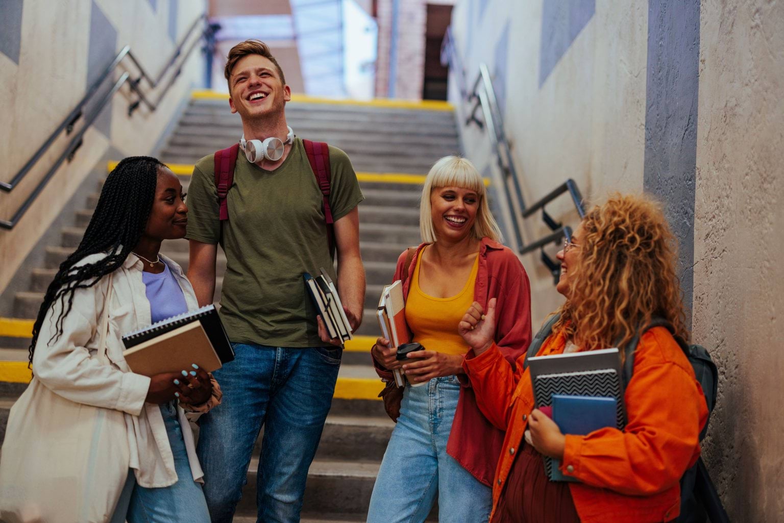 Four students smiling and holding books, standing at the bottom of a staircase