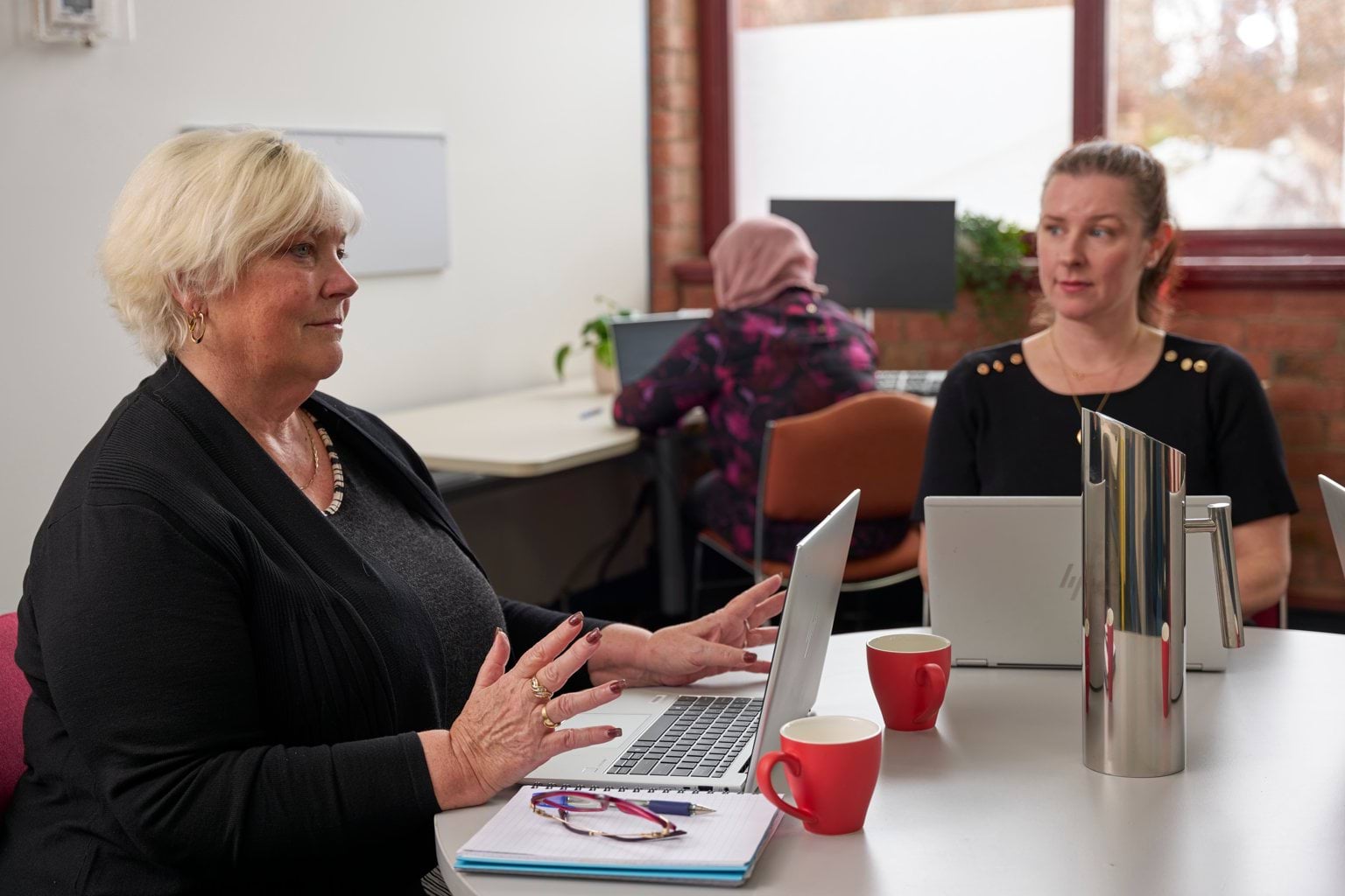 Two people sit at a table with computers, having a discussion.