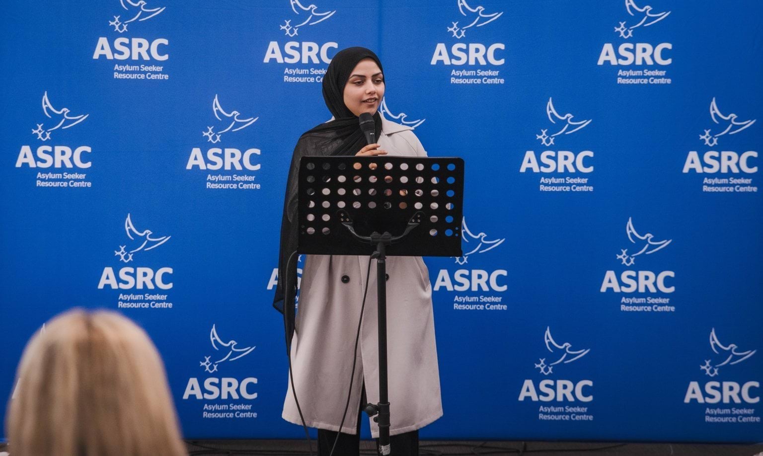 A person speaking at a podium with a blue backdrop that has the Asylum Seeker Resource Centre logo repeated.