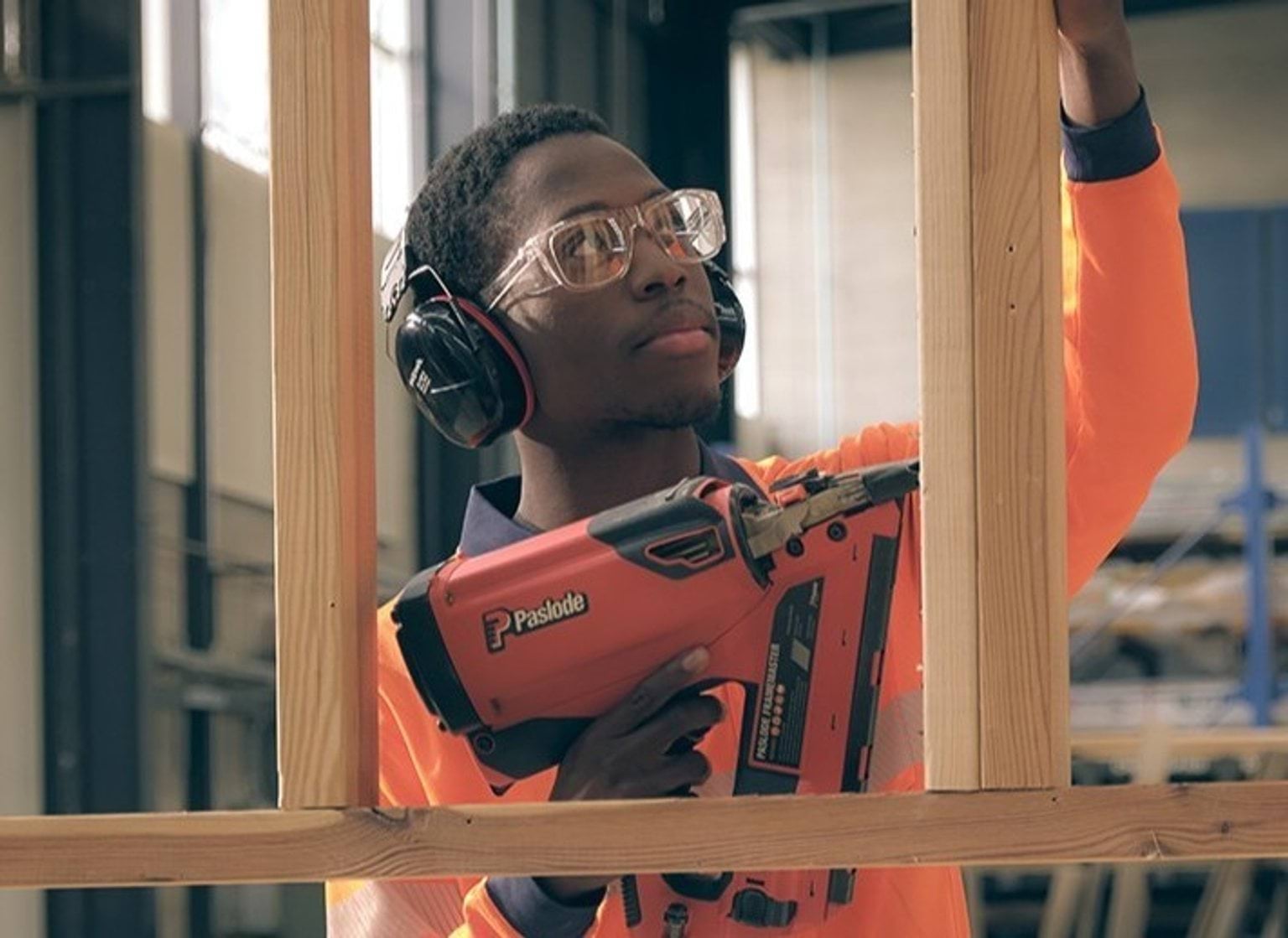 A person in hi-vis, headphones and safety glasses uses a nail gun on a wooden frame.