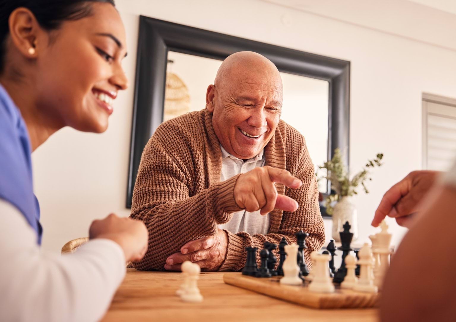 An older person is playing chess. A person in medical scrubs sits next to him.