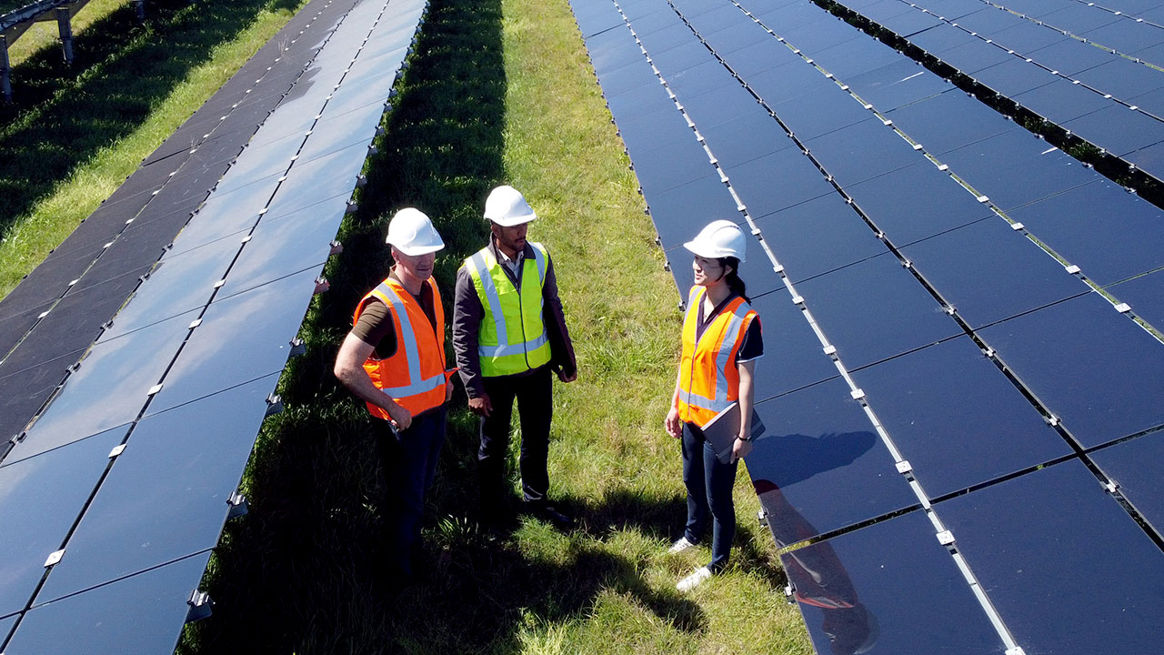 Three people standing between long rows of solar panels wearing high vis vests and safety helmets