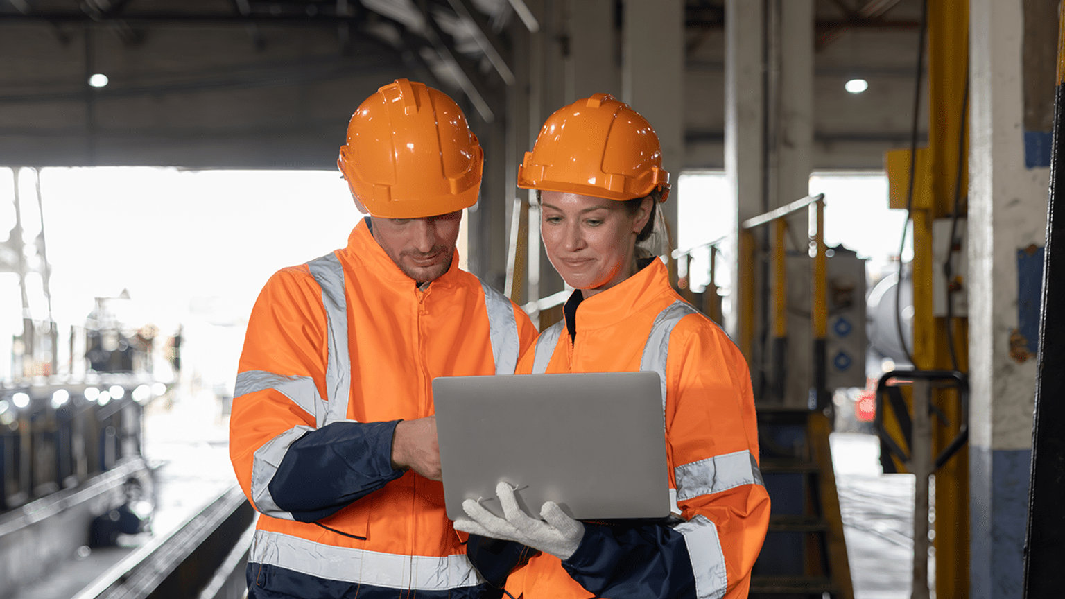 Two people in hard hats using a laptop in an industrial setting.