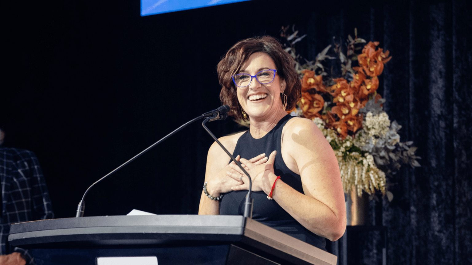 A person at a podium smiles and holds their hands to their chest excitedly.