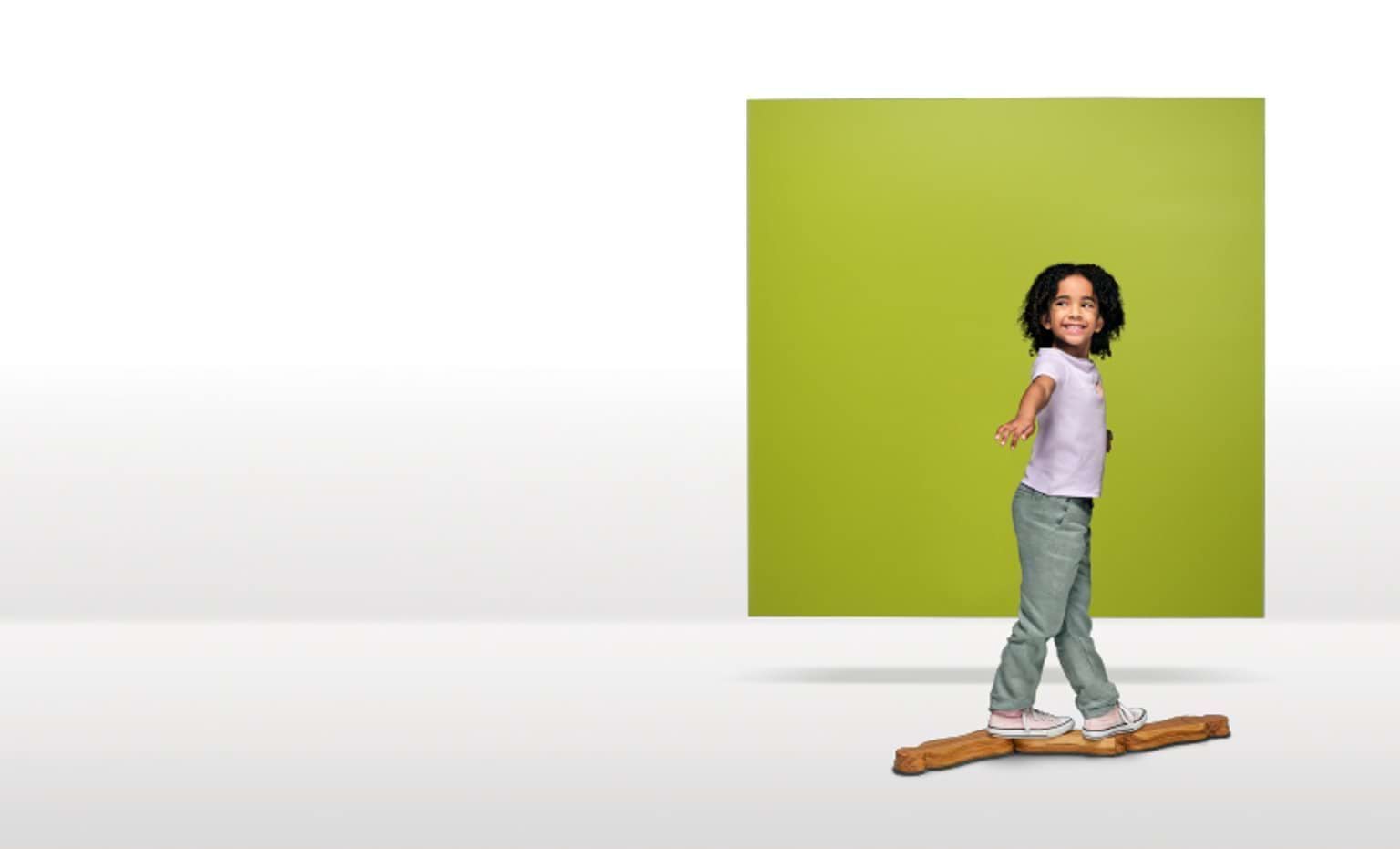 A 3-year-old child stands on balance beam in front of a green square floating in a white space.