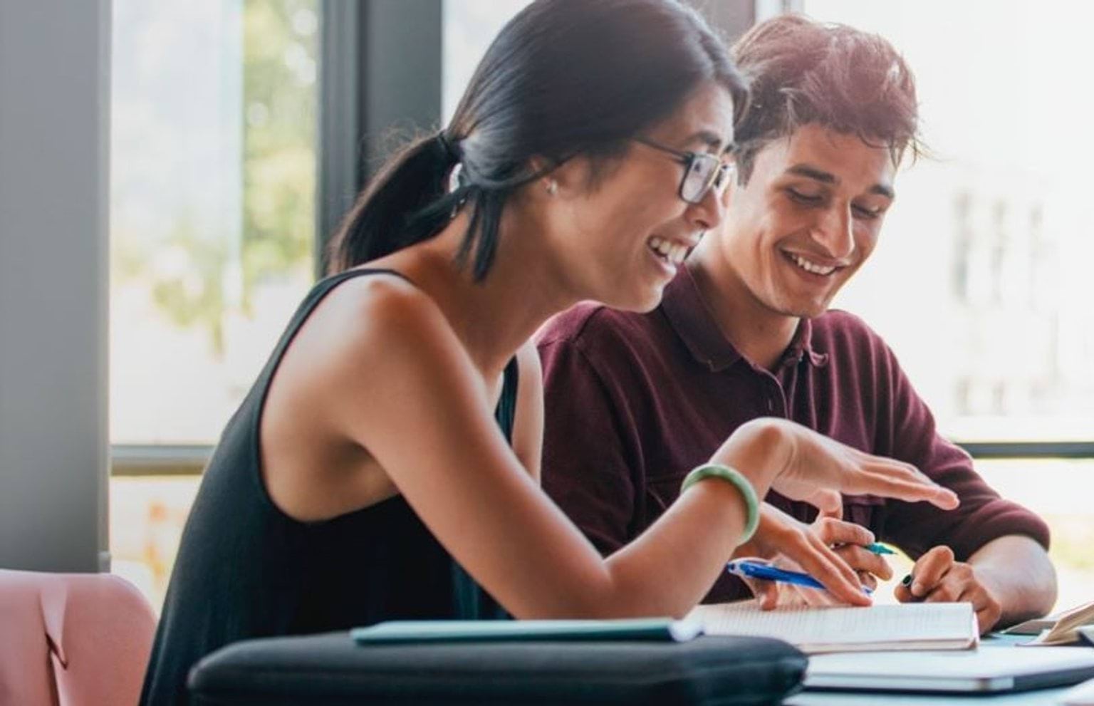 Two students sitting at a desk, holding pens and smiling.